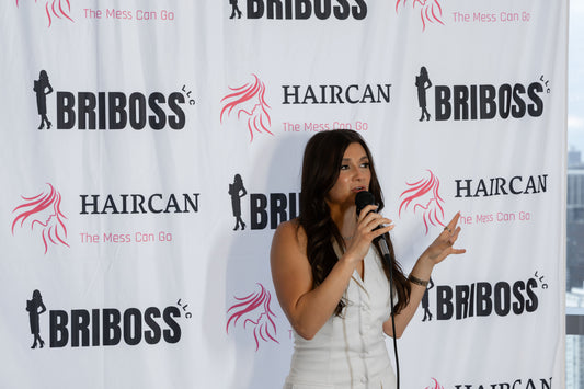 Woman speaking into a microphone at an event with Briboss and Haircan branding in the background. 1